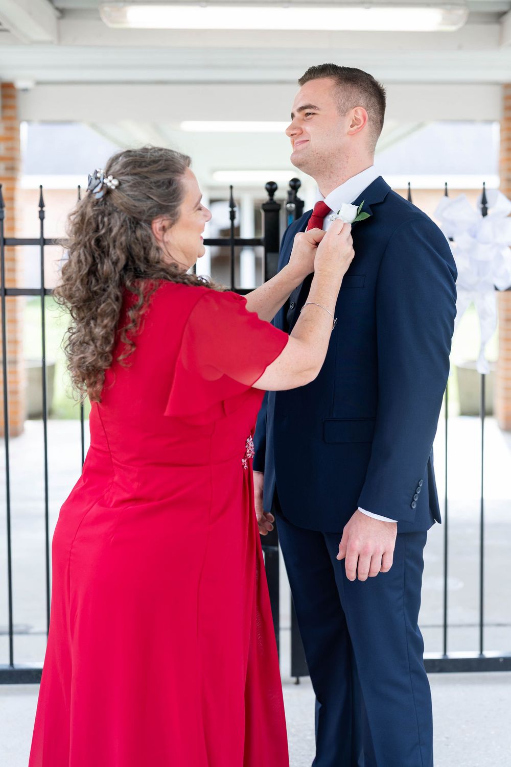 Woman in red dress, mom, adjusting groom's red tie before wedding ceremony outdoors.