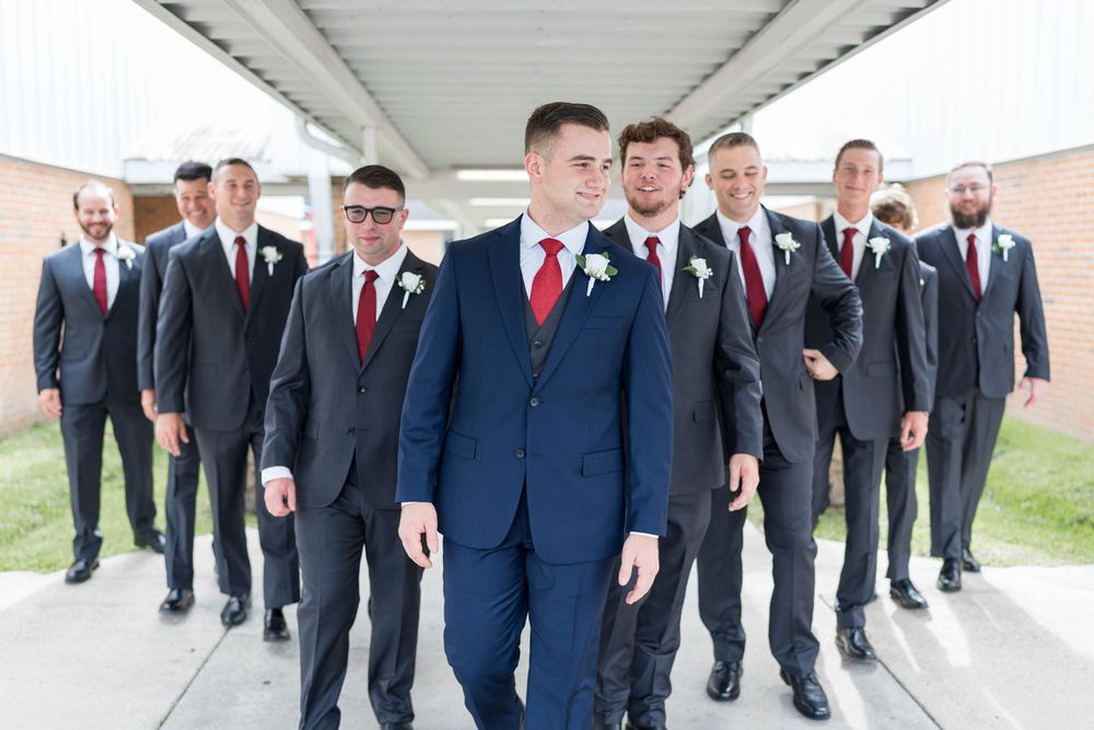 Groom in navy suit leads groomsmen in gray suits with red ties walking outdoors at a wedding.