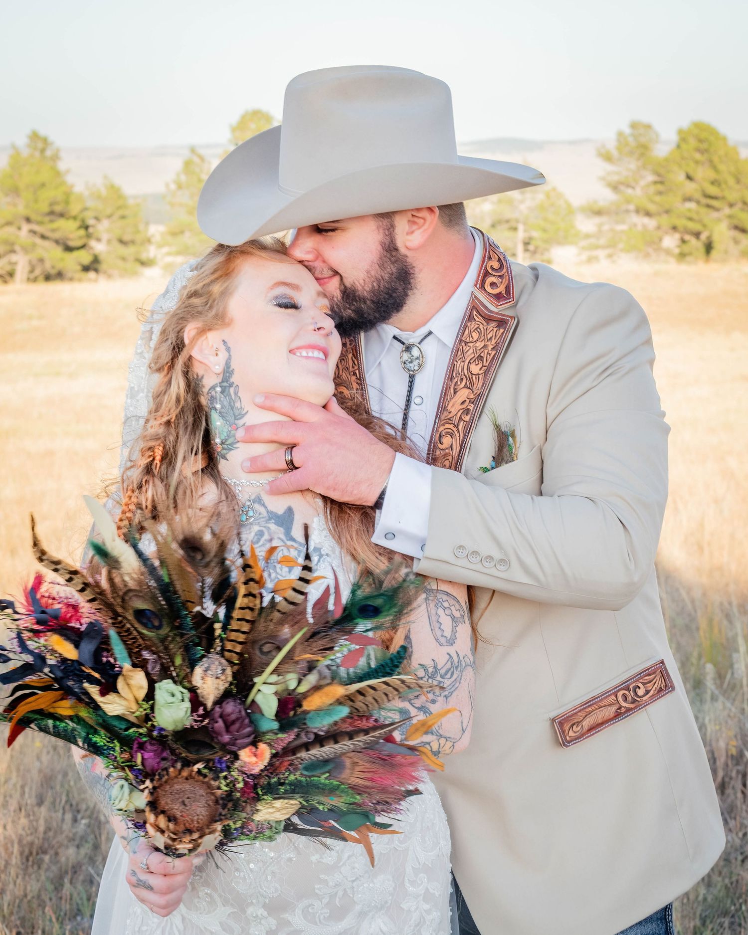 Couple in western wedding attire embrace in golden field, groom in cowboy hat kisses bride holding colorful feather bouquet.