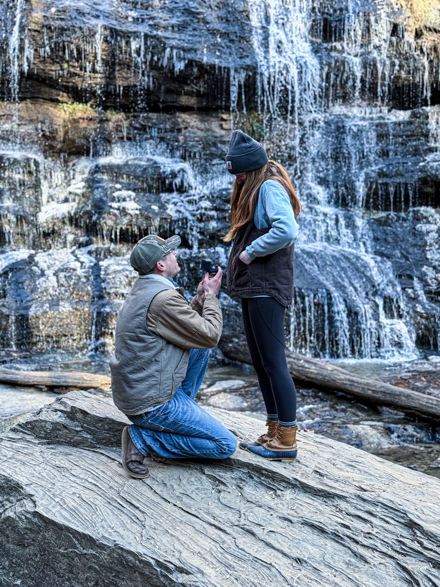Man proposes on one knee to woman in front of stunning waterfall with icicles on rocky ledge outdoors.