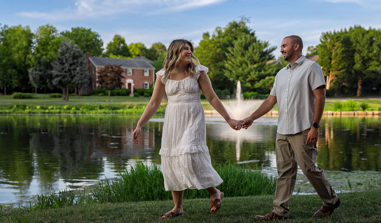 Couple walking hand-in-hand on a scenic path by a pond with a red brick building and lush green trees at sunset.