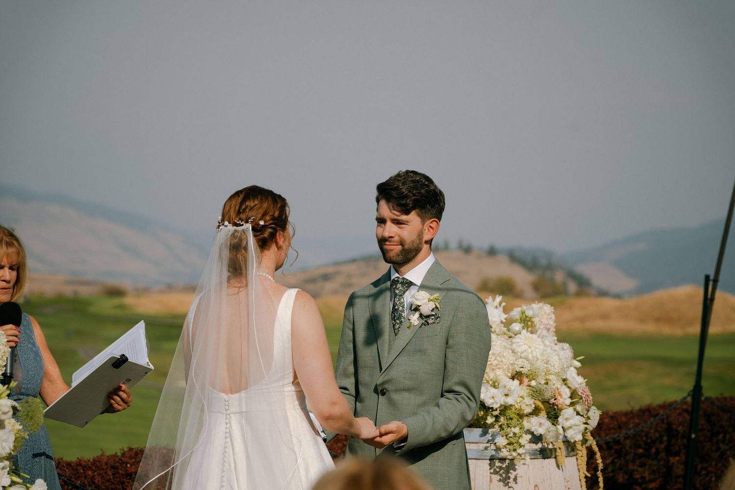 Bride and groom exchanging vows at outdoor wedding ceremony with rolling hills and floral arch backdrop.