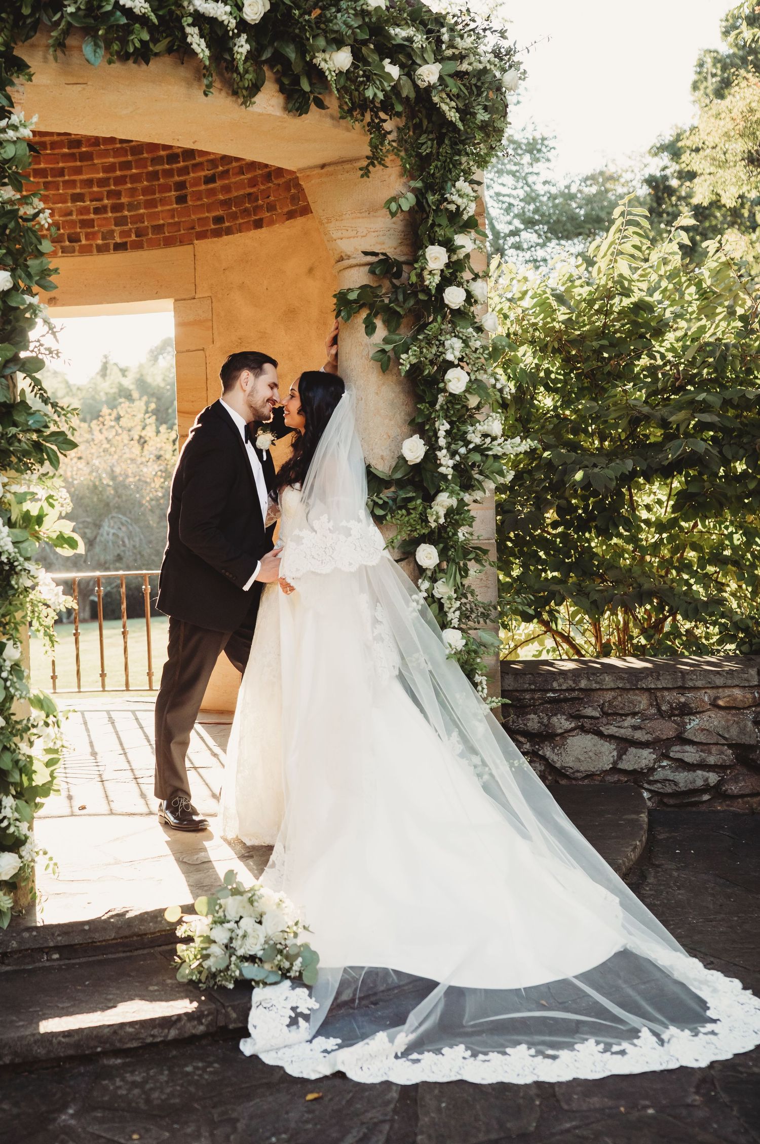 Bride and groom share romantic moment under floral arch with white roses and greenery at outdoor wedding ceremony.