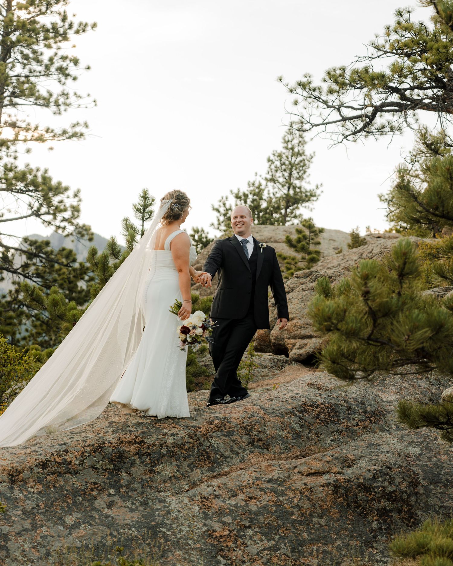 A bride and groom hold hands on a rocky mountain overlook surrounded by pine trees during their outdoor wedding ceremony.