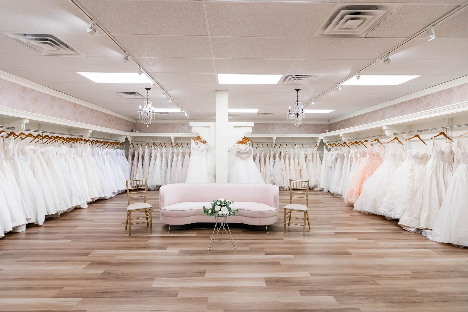 Elegant bridal boutique showroom with rows of white wedding dresses displayed on racks and a pink velvet sofa centerpiece.