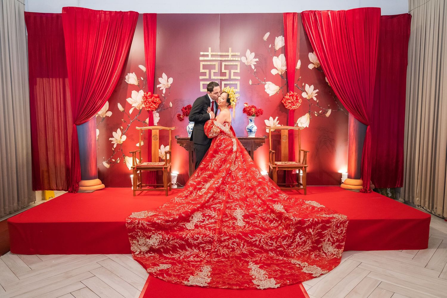 Couple in traditional Chinese wedding attire on red stage with floral backdrop and double happiness symbol.