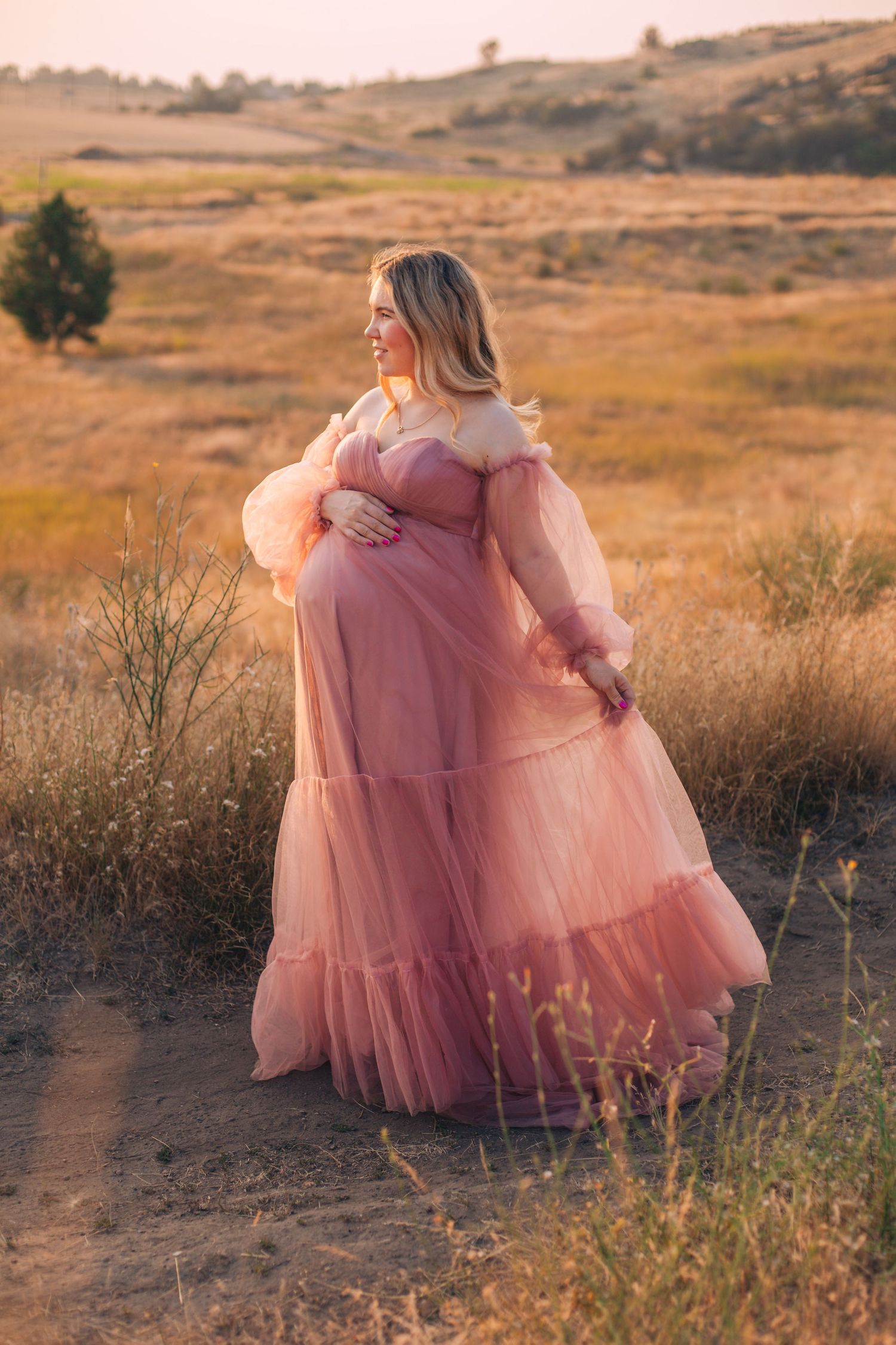Pregnant woman in pink tulle maternity gown posing in golden field at sunset.