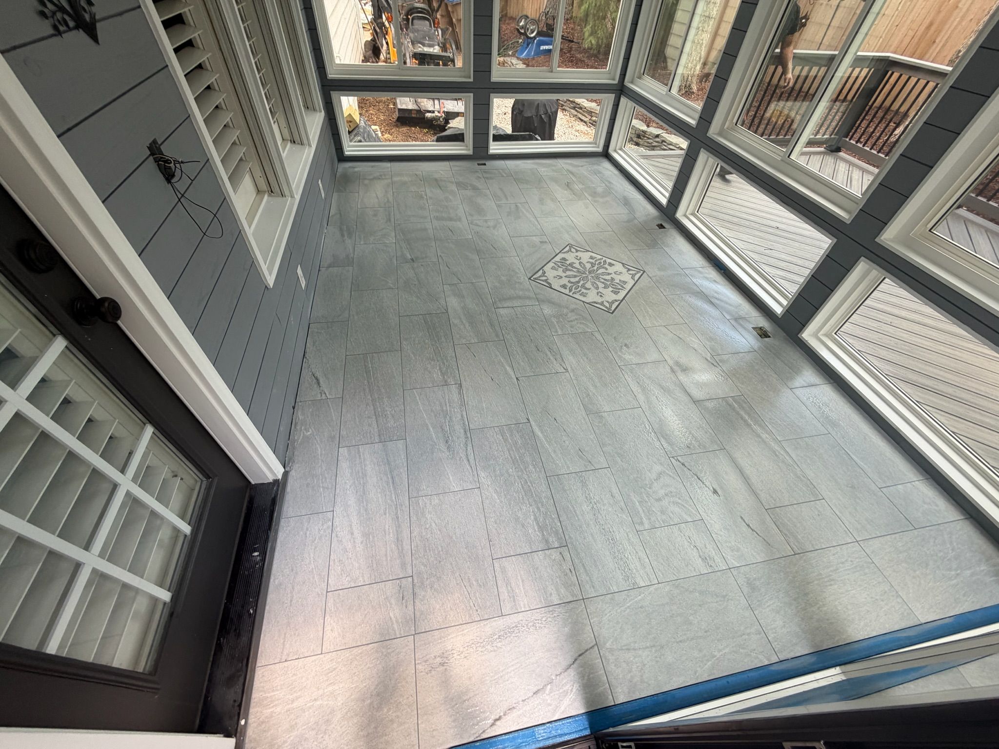 Gray tile flooring installation in a sunroom with decorative mosaic center medallion and large glass windows.