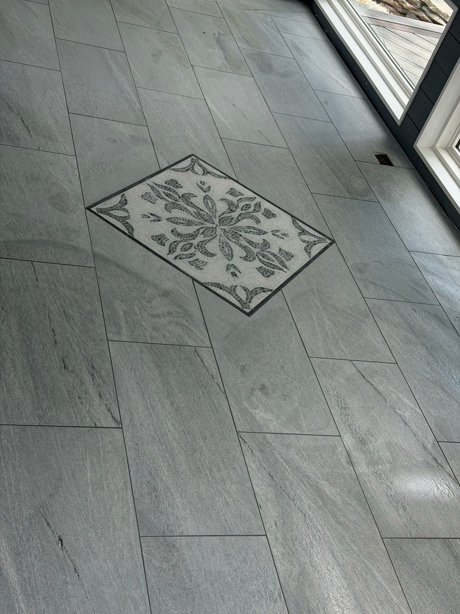 Gray stone-look tile floor with a decorative rectangular floral mosaic inlay accent piece near glass door.