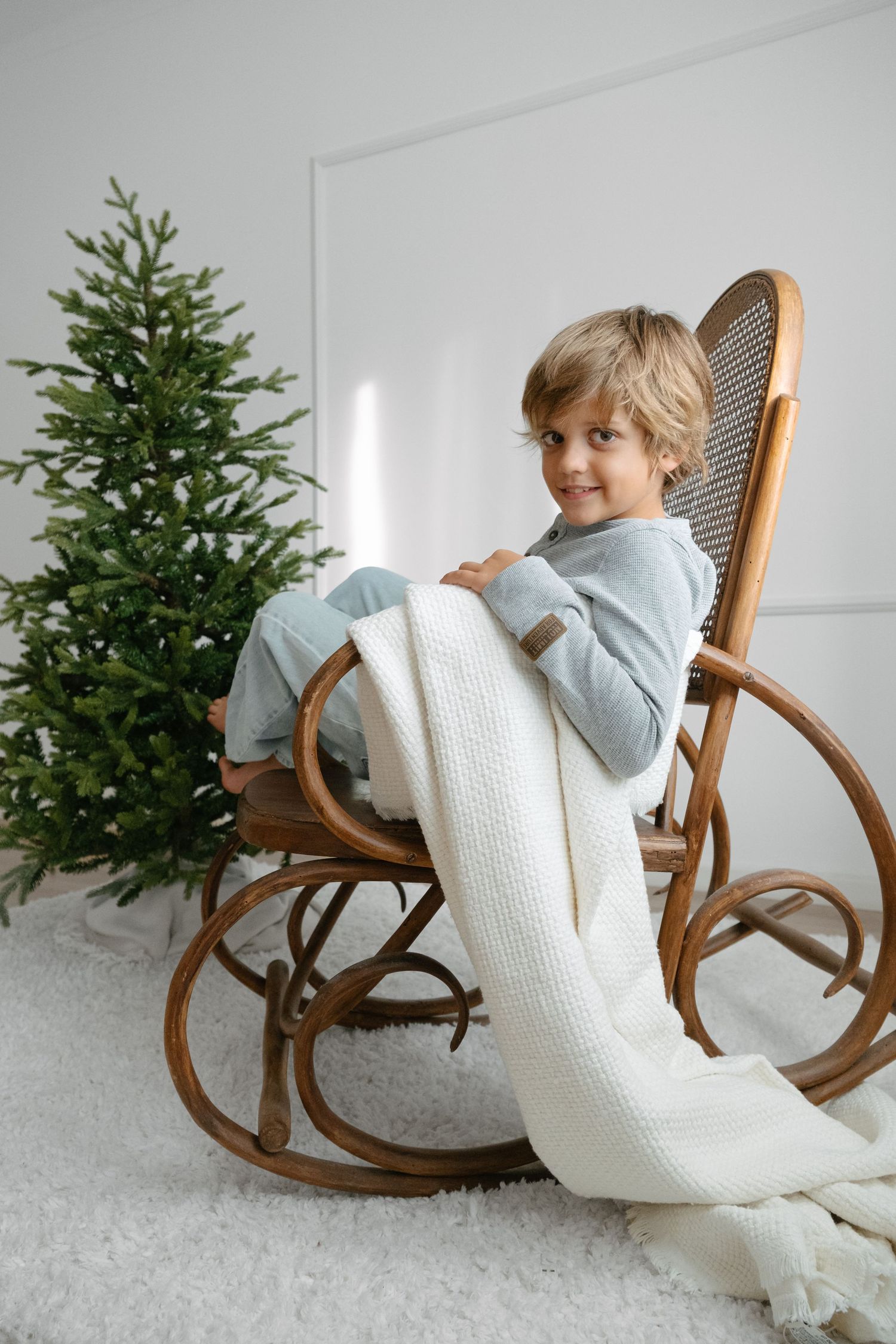 Young boy sitting in wooden rocking chair with white knit blanket beside decorated Christmas tree.