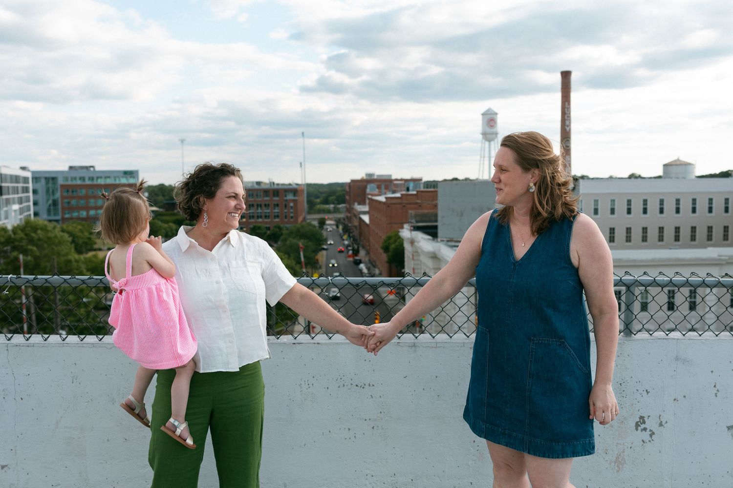 Family of three on rooftop parking lot, parents with toddler girl in pink dress, city skyline background.