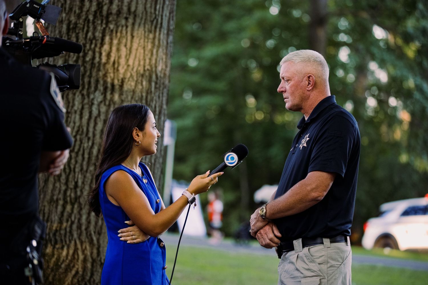 Female reporter interviews a law enforcement officer outdoors near a tree during a news segment.