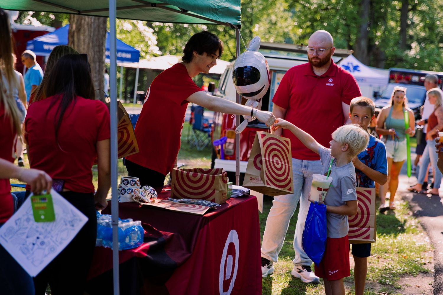 Target mascot interacts with children at an outdoor community event booth staffed by employees in red shirts.