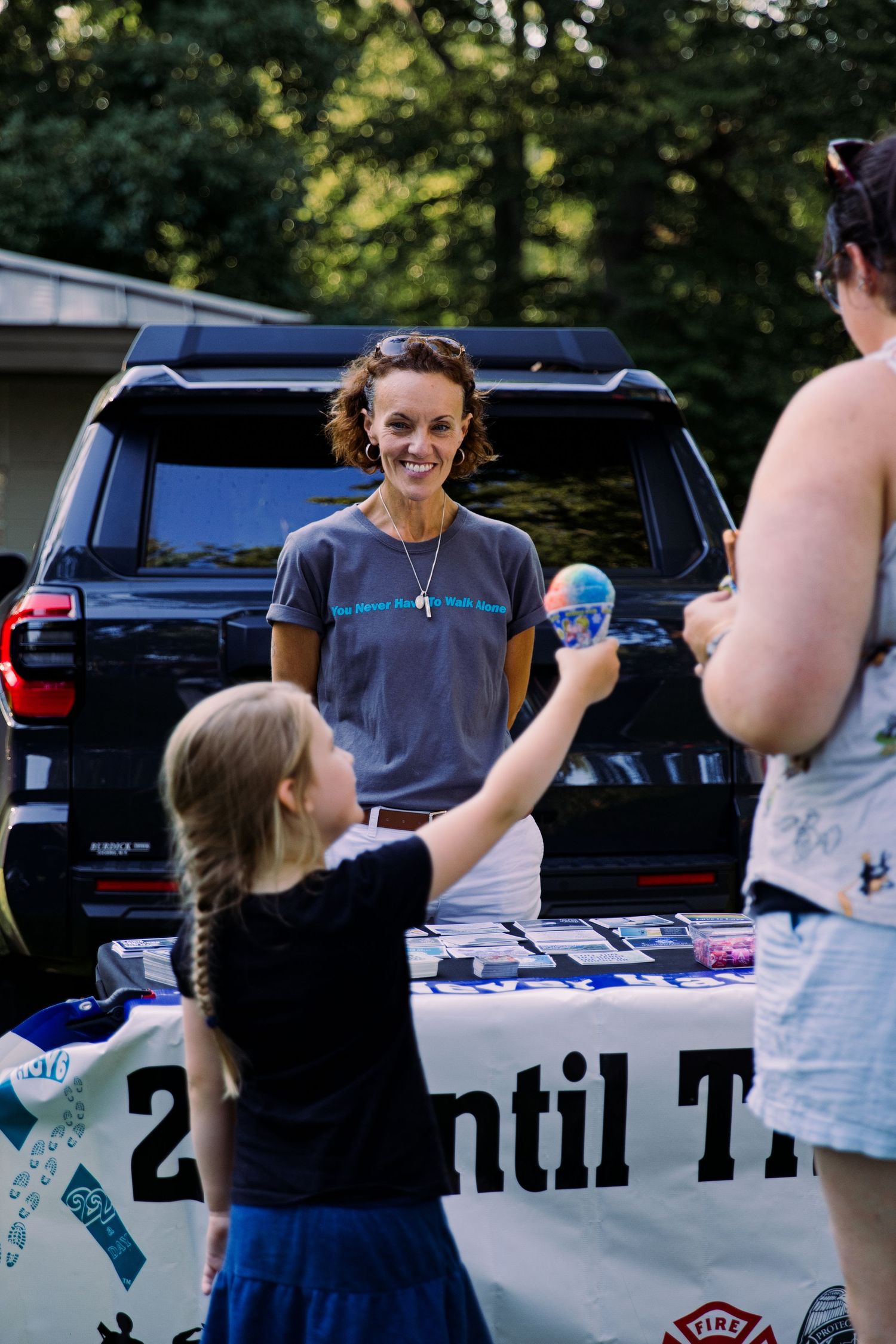 Woman in gray t-shirt smiles at a child reaching up at an outdoor community event table with a banner.