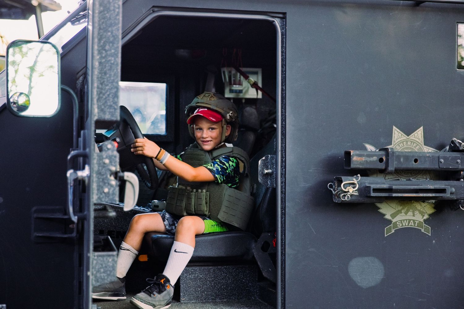 Young boy in military gear sitting in driver seat of armored police vehicle, smiling at camera.