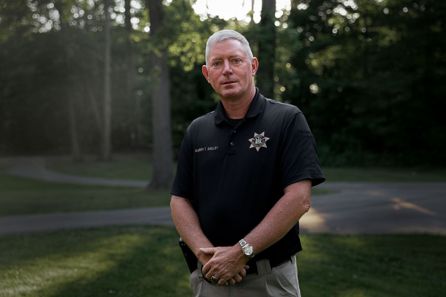 A law enforcement officer in a black polo shirt with a badge stands outdoors in a park at dusk.