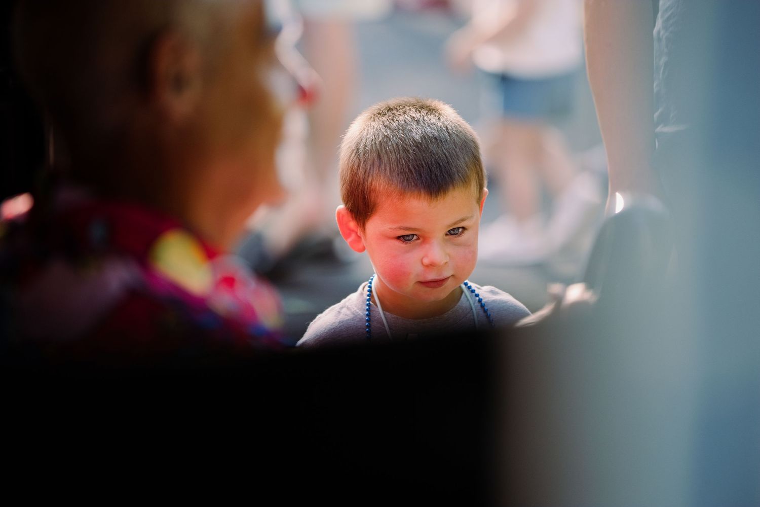 Young boy with short brown hair looking thoughtful at an outdoor gathering with adults nearby.