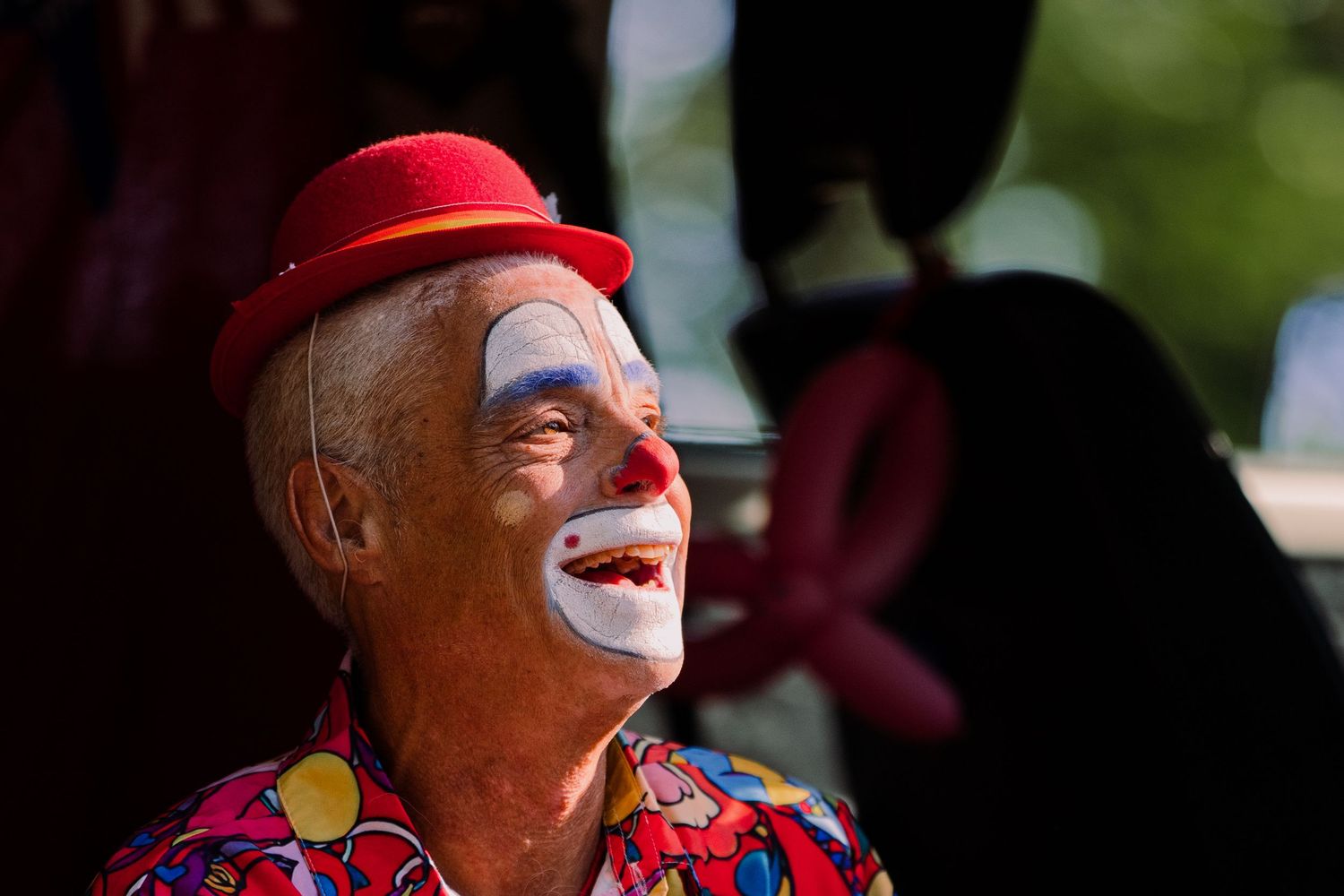 Elderly clown with white face paint and red hat smiling outdoors in colorful costume.