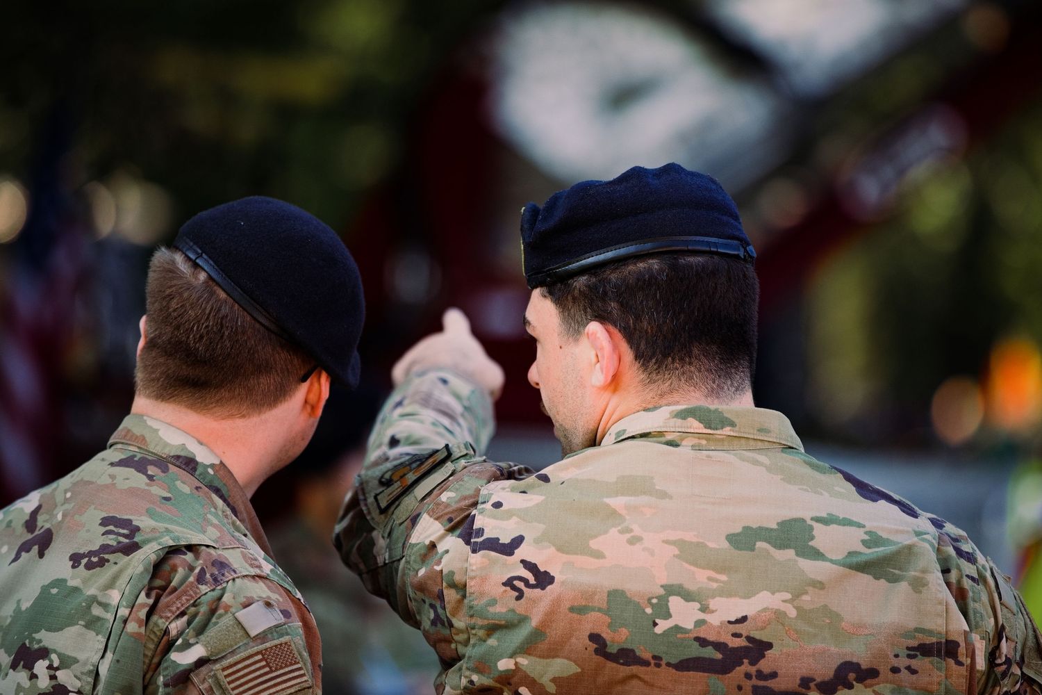 Two military soldiers in camouflage uniforms and berets pointing and discussing during an outdoor ceremony.