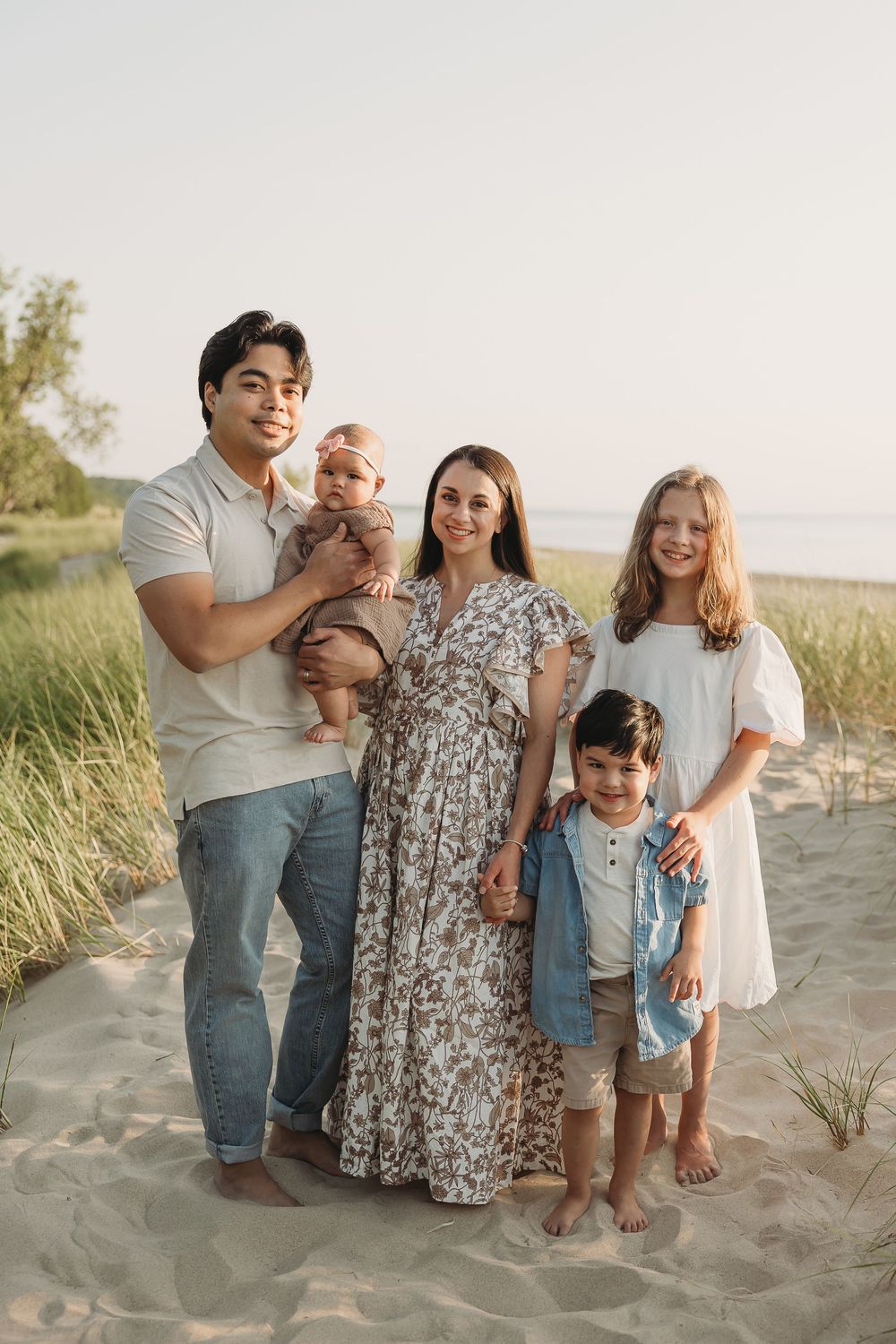 Happy family of five posing on sandy beach at sunset, parents with three children including baby.
