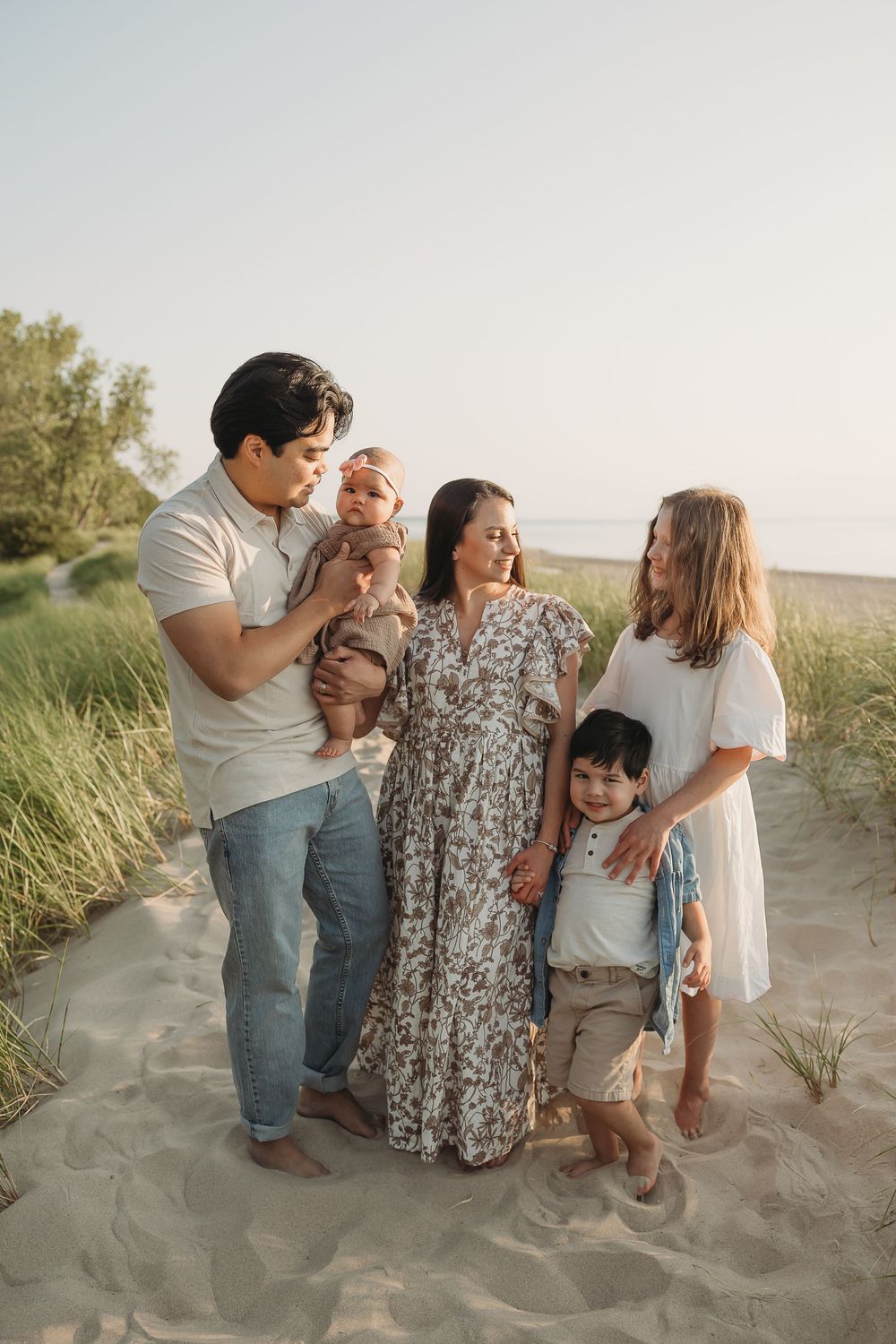 Family of five posing on sandy beach dunes at golden hour, parents with three children in casual neutral outfits.