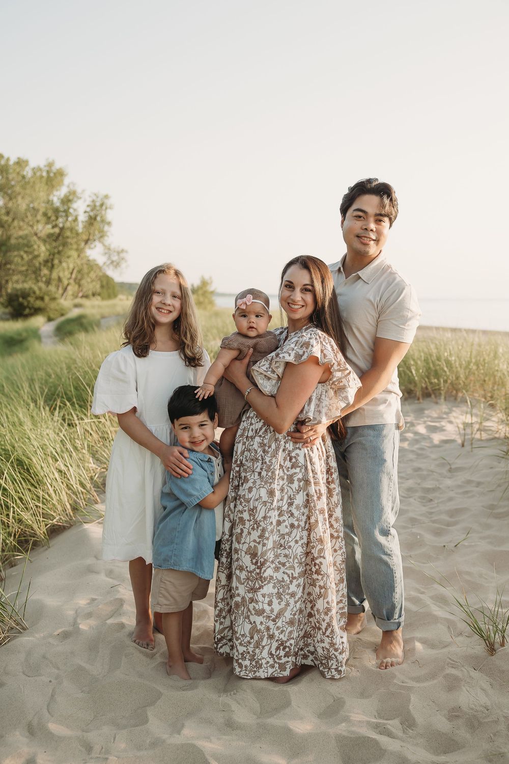 Family of five posing on sandy beach dunes, parents with three children smiling outdoors in casual summer attire.