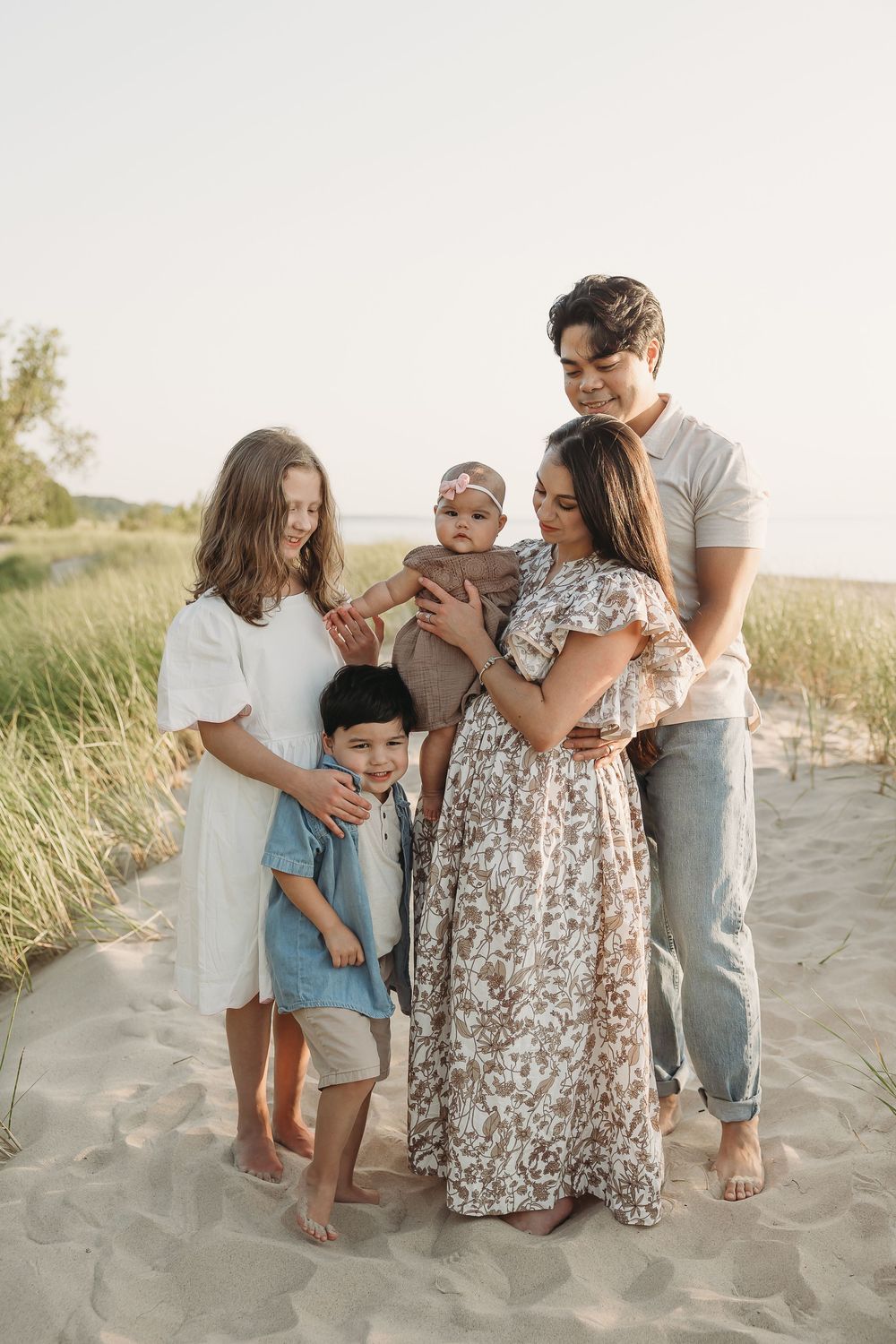 A happy family of five poses together on a sandy beach path surrounded by dune grass in soft natural light.