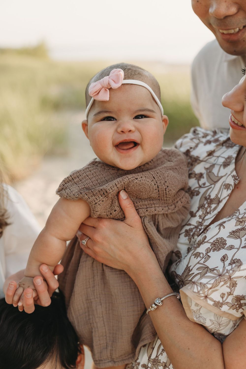Smiling baby girl in brown dress and pink bow headband being held by parents outdoors.