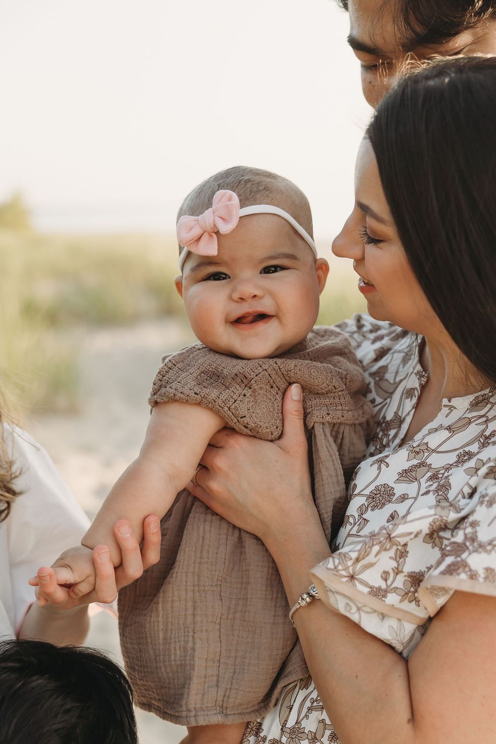 A smiling baby girl in a brown dress and pink bow headband being held by her mother outdoors.