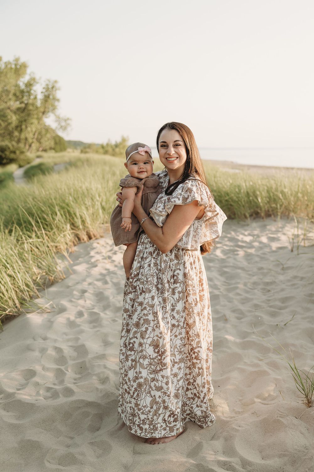 Smiling mother in floral maxi dress holds baby on sandy beach path with green dunes in background.