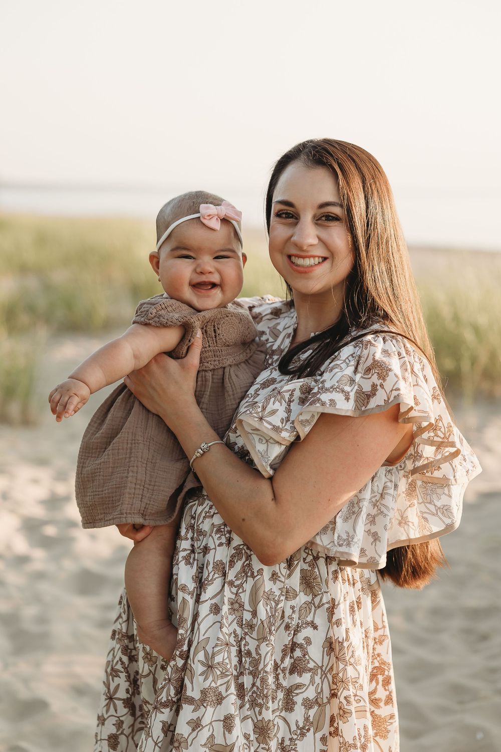 Smiling mother holding happy baby girl on sandy beach, both wearing floral and brown outfits.