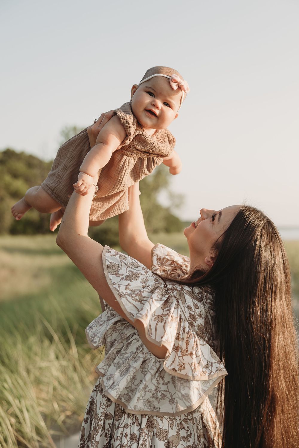 A mother lifts her smiling baby girl in the air outdoors, both wearing matching floral dresses in a sunny field.