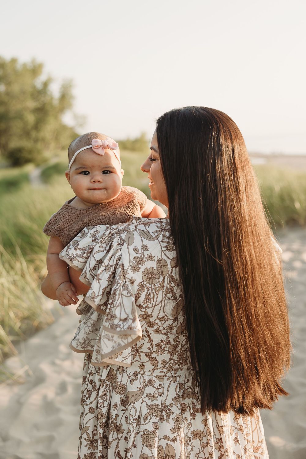 A mother with long dark hair holds her baby girl on a sandy beach path, both dressed in warm tones.