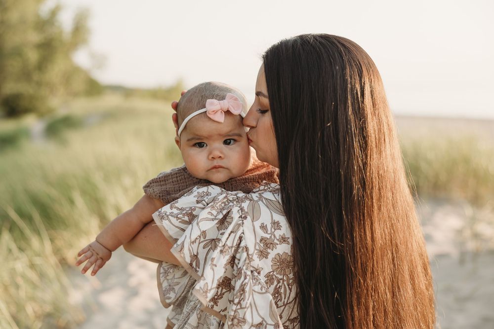 A mother with long dark hair kissing her baby girl wearing a pink headbow on a sandy beach.