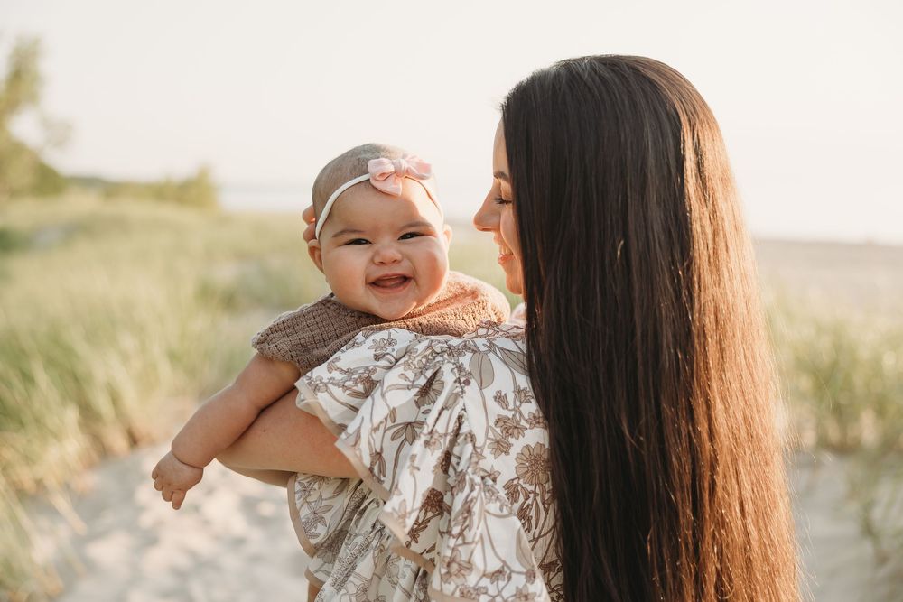 A smiling baby girl with a pink bow headband being held by her long-haired mother on a sunny beach.