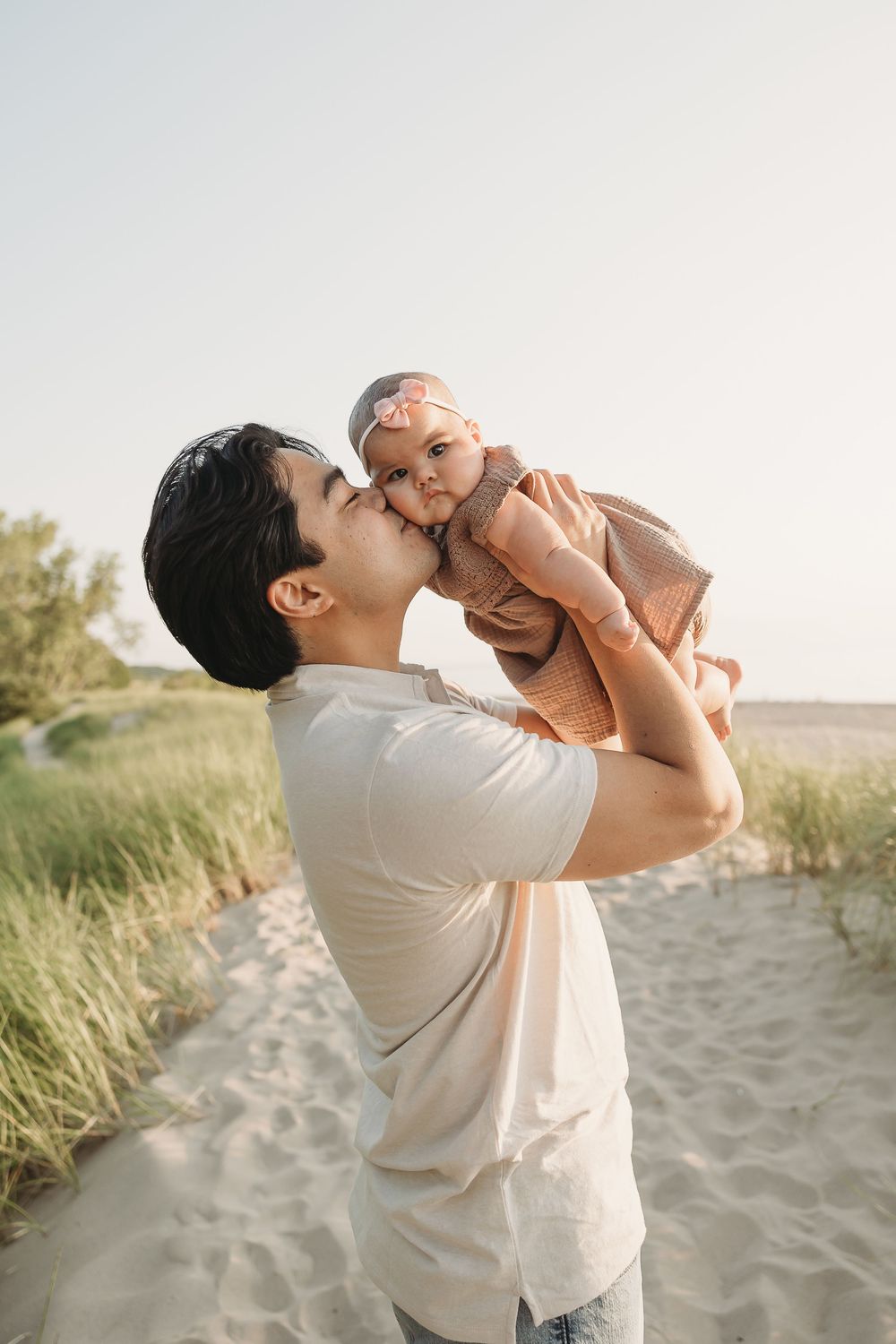 A father lovingly kisses his baby girl while holding her up at a sandy beach.