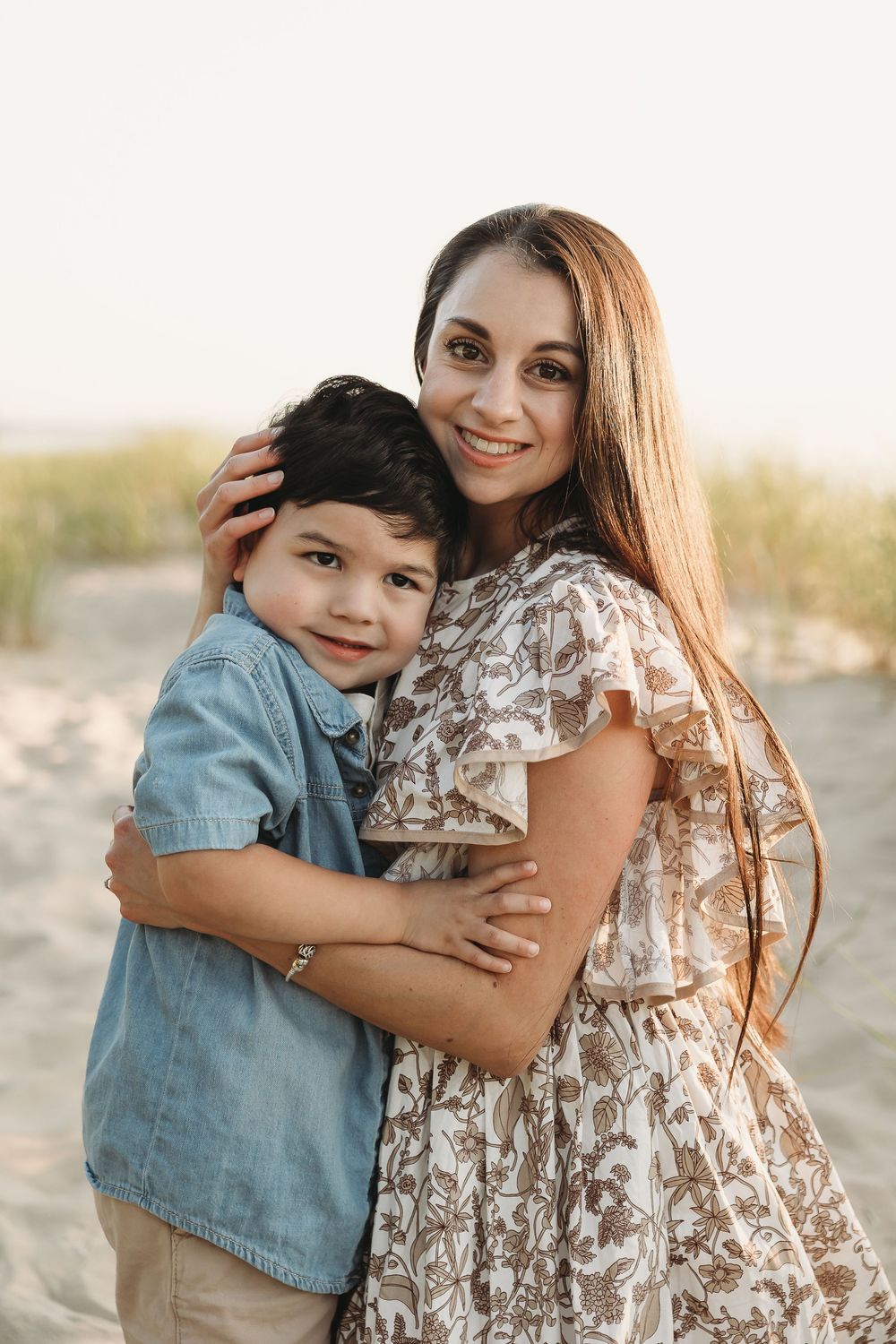 A woman in a floral dress hugs a young boy in a denim shirt on a sandy beach at sunset.