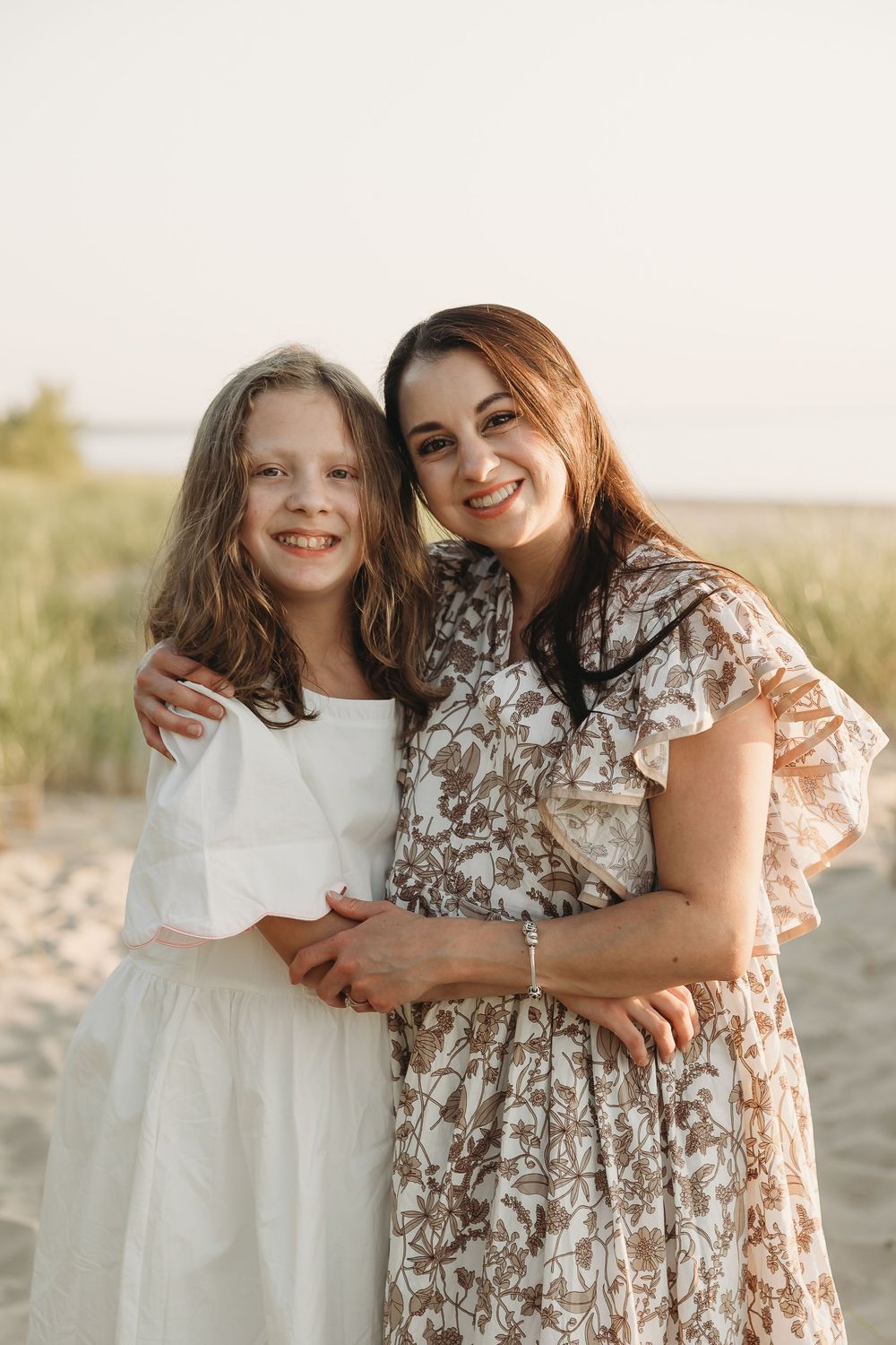 A woman and young girl in dresses smile together on a sandy beach in warm natural light.