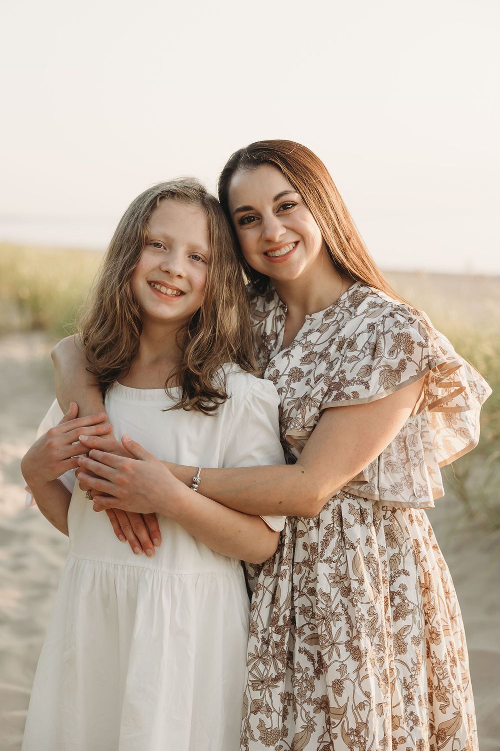 A woman in a floral dress hugs a smiling girl in white on a sunny beach.