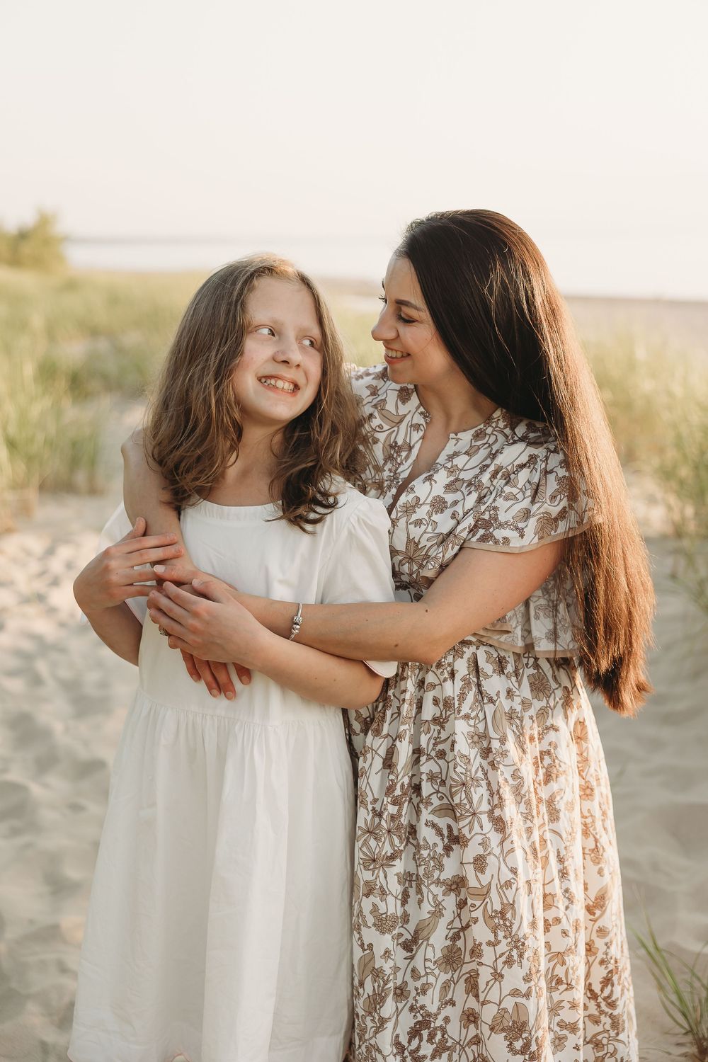 A mother and daughter embrace on a sandy beach at sunset, wearing floral and white dresses, smiling warmly at each other.