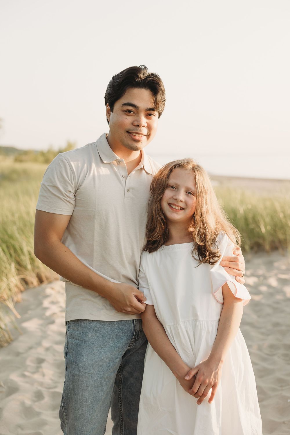 A young man and girl smiling together on a sandy beach, wearing casual white outfits in warm golden light.