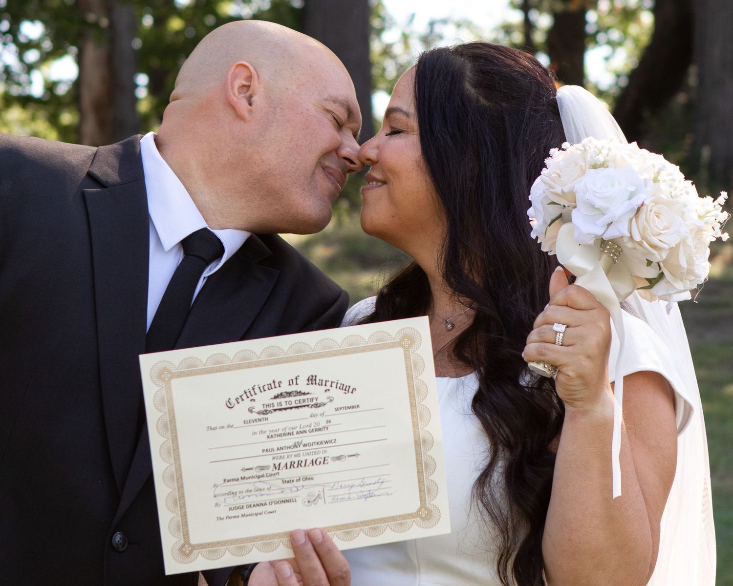 Newlywed couple kissing while holding marriage certificate and white bouquet in wedding attire outdoors.