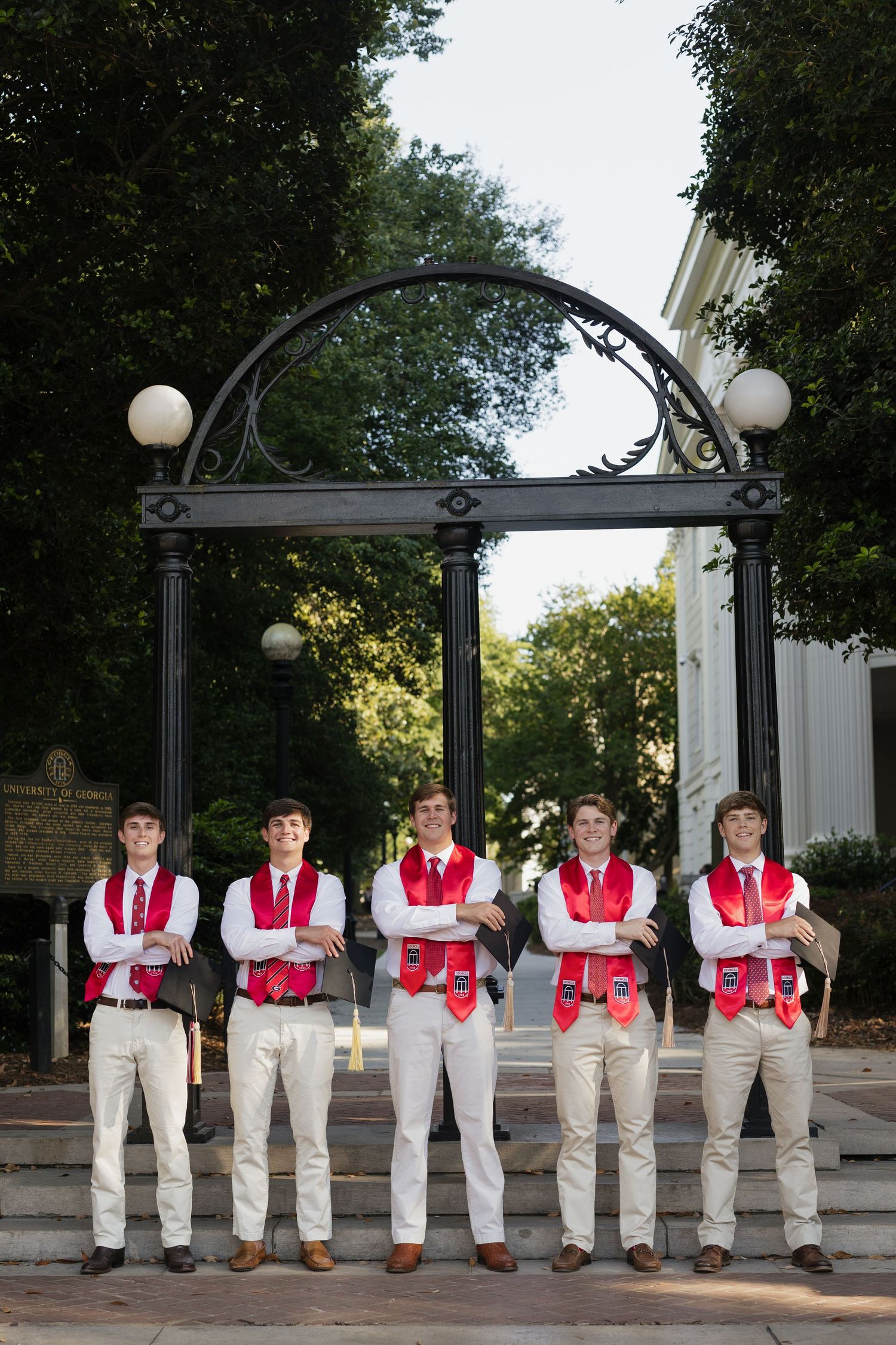 Five young men in matching red vests and ties pose with arms crossed under an ornate arch on college campus.