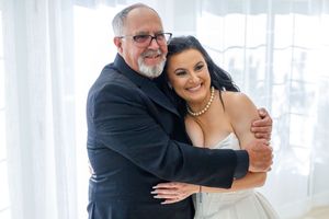 A bride in a white gown and pearl necklace hugs an older man in a dark suit, smiling warmly near a bright window.