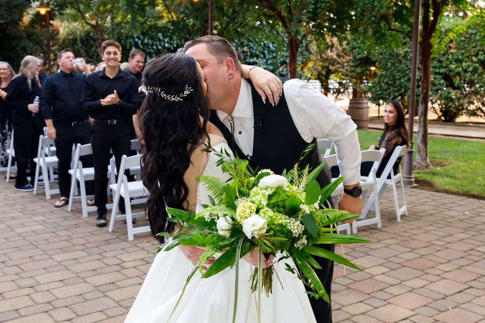 Bride and groom share a kiss at outdoor wedding ceremony with white chairs and guests in background.