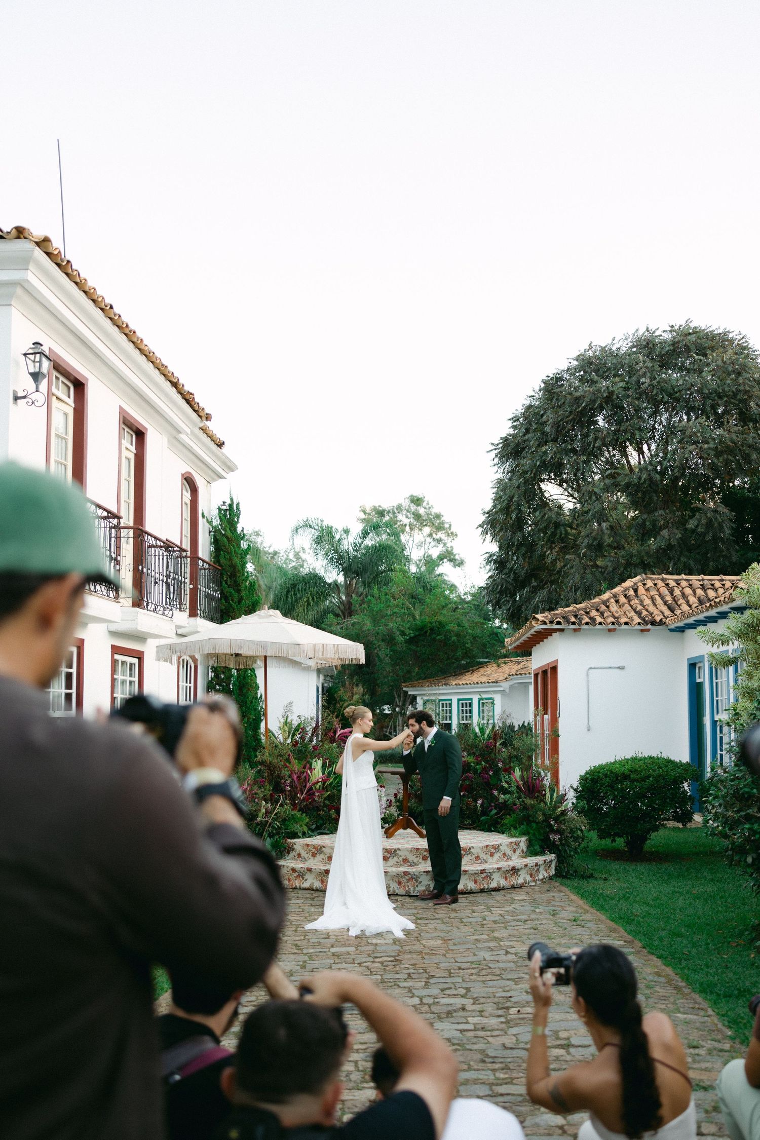 Bride and groom share a romantic moment outdoors at a colonial-style venue while photographers capture the wedding ceremony.