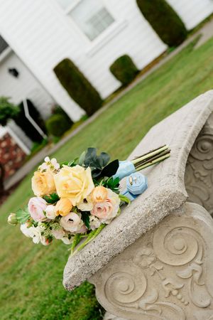 Bridal bouquet of yellow and pink roses with a blue ring box resting on an ornate stone bench in a garden.