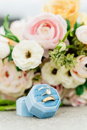 Gold wedding rings in a blue velvet ring box with a pastel floral bouquet of roses and ranunculus in the background.