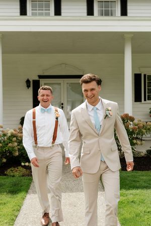 Two grooms in tan suits with blue ties walk happily toward camera outside a white wedding venue.