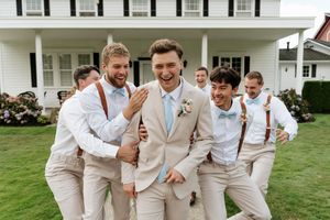 Groom in tan suit surrounded by laughing groomsmen in white shirts and suspenders outside a white house.